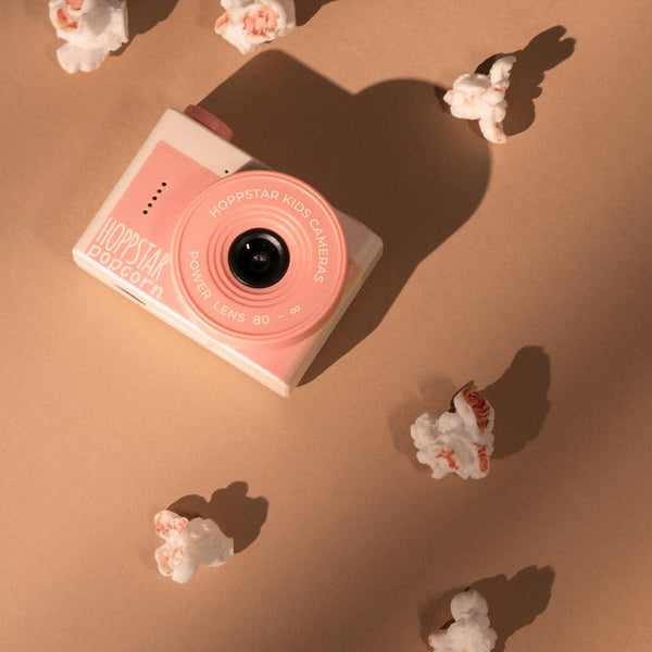 A compact instant camera in pastel pink and white lies on a warm tan surface with soft diagonal shadows and small white flower props around it.